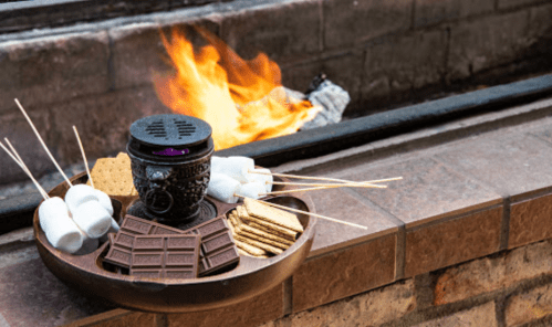 A tray with graham crackers, chocolate, marshmallows, and a small fire pit in the background, ready for s'mores.