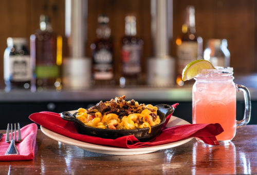 A skillet of cheesy macaroni with pulled pork, served with a pink drink in a mason jar, on a wooden table.