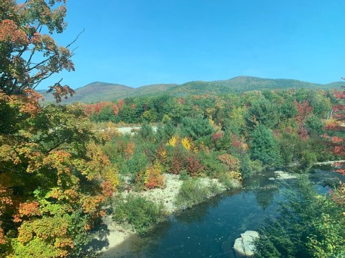 A vibrant landscape featuring colorful autumn foliage along a river, with mountains in the background under a clear blue sky.