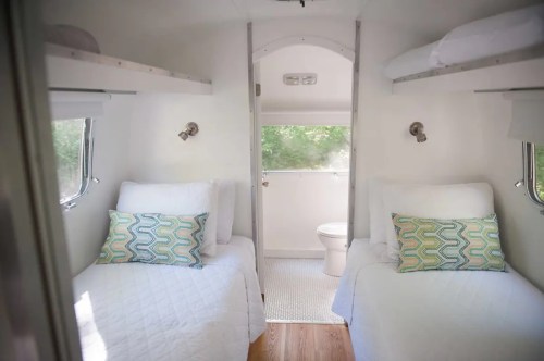 Cozy interior of a small trailer with two twin beds, colorful pillows, and a bathroom visible in the background.