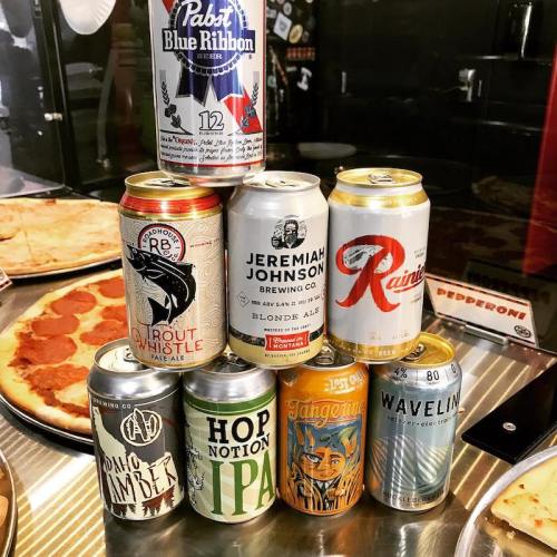 A stack of various beer cans on a table with pizza in the background.