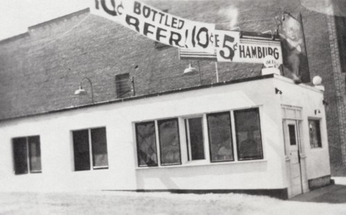 Black and white photo of a small building with signs advertising bottled beer and hamburgers.