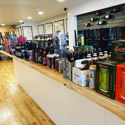 A bar counter lined with various beer cans and bottles, with empty chairs and framed artwork in the background.