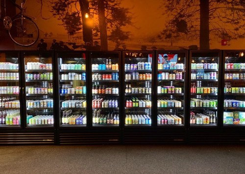 A well-stocked beverage cooler with various colorful cans and bottles, illuminated in a dimly lit store.