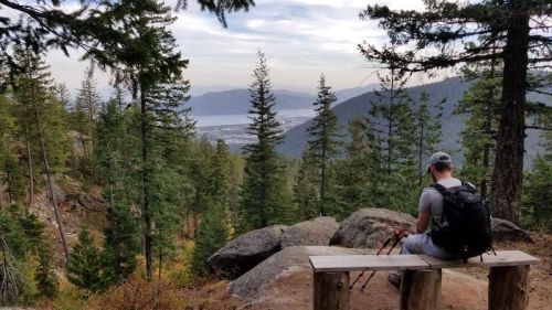 A person sits on a bench in a forest, overlooking a valley and lake surrounded by mountains and trees.