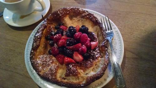 A golden-brown pancake topped with a variety of fresh berries and powdered sugar, served on a plate with a fork.