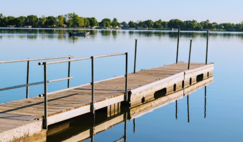 A wooden dock extends over a calm lake, reflecting trees and a clear blue sky. A small boat is visible in the distance.