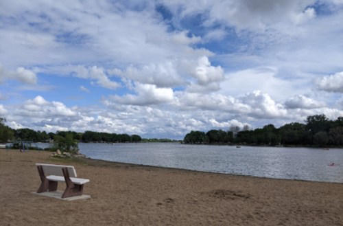 A sandy beach by a calm lake, with a bench and fluffy clouds in a blue sky.