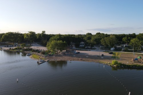 Aerial view of a serene lake with a sandy beach, trees, and RVs parked nearby under a clear blue sky.