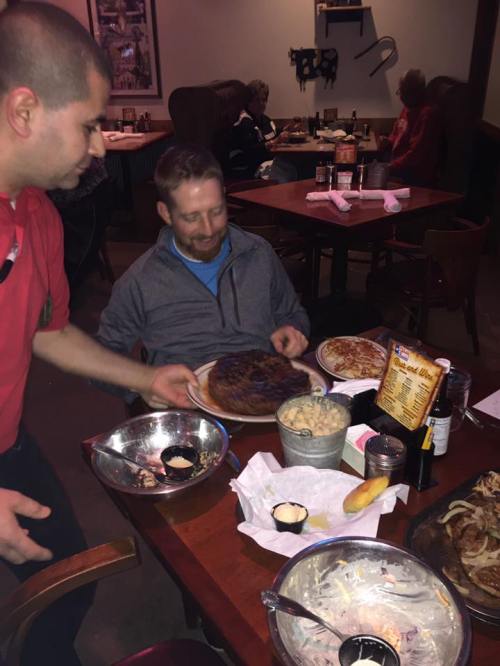 A waiter serves a large plate of food to a smiling man at a restaurant table, surrounded by other diners.