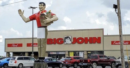 A large statue of a man holding groceries stands in front of a store named "Big John." Cars are parked nearby.