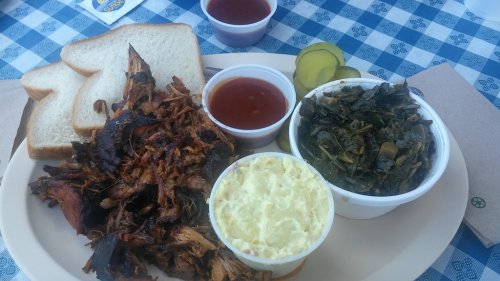 A plate of pulled pork with barbecue sauce, sides of collard greens and potato salad, and slices of bread.