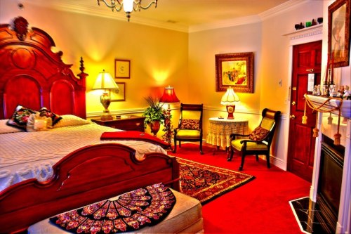 Cozy bedroom with a wooden bed, decorative lamps, a small table, and a fireplace, featuring warm lighting and rich colors.