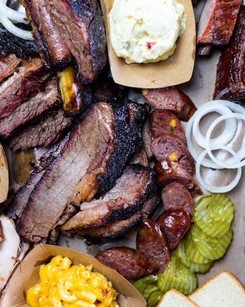 A close-up of a barbecue platter featuring sliced brisket, sausage, pickles, onions, macaroni and cheese, and bread.