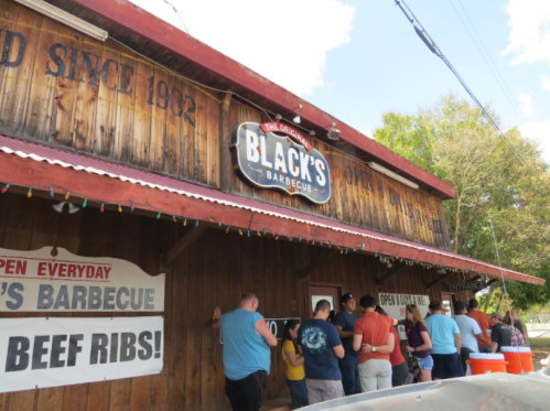 A crowd lines up outside Black's Barbecue, a rustic wooden building with signs advertising beef ribs and open hours.