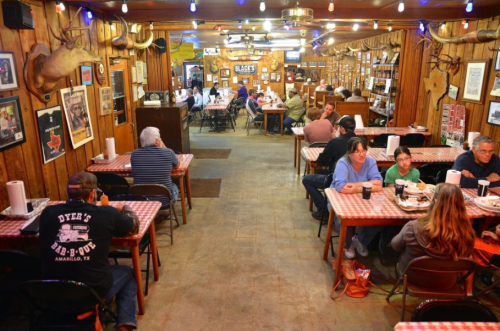 A bustling barbecue restaurant with wooden decor, checkered tablecloths, and diners enjoying meals at various tables.