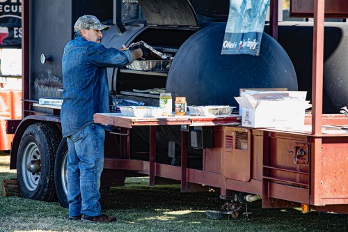 A man in a denim jacket prepares food at a large outdoor grill on a trailer at a community event.