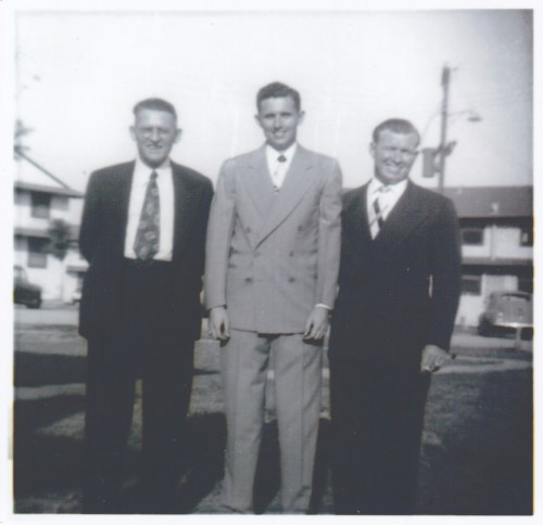 Three men in suits stand together outdoors, smiling, with houses and a car visible in the background.