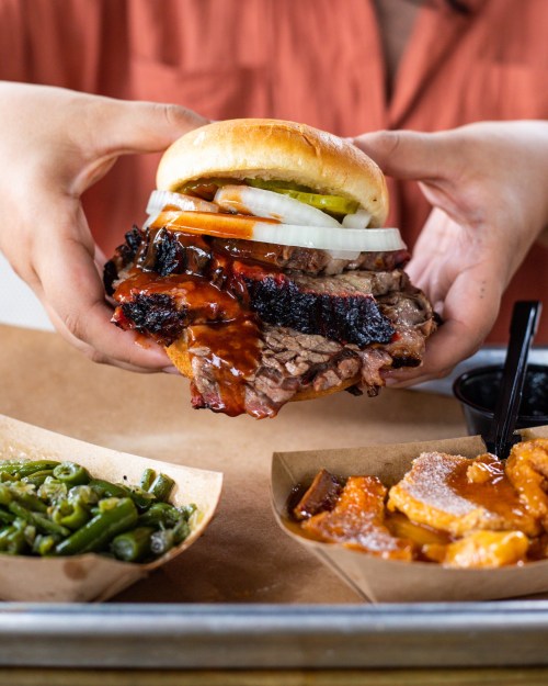 A person holds a large sandwich filled with brisket, pickles, onions, and barbecue sauce, with sides of green beans and sweet potatoes.