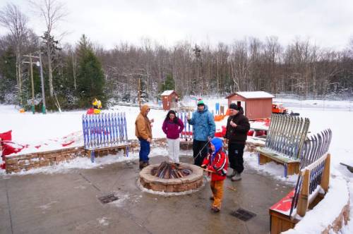 A group of people in winter clothing gathered around a fire pit in a snowy outdoor setting.