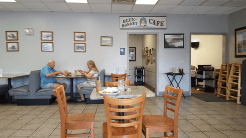 A cozy café interior with two patrons at a table, framed pictures on the walls, and a view of the kitchen area.