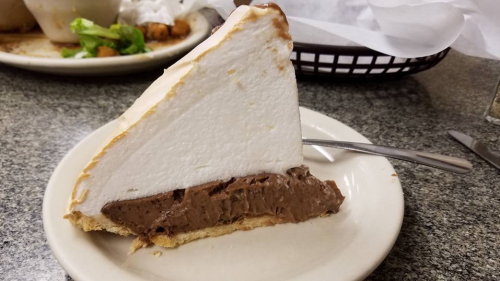 A slice of chocolate cream pie with a fluffy meringue topping on a white plate, set on a gray table.