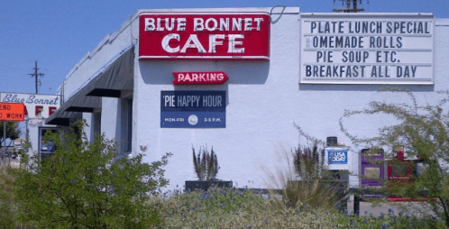 Signage for Blue Bonnet Café featuring "Pie Happy Hour," daily specials, and parking information. Desert landscape in foreground.