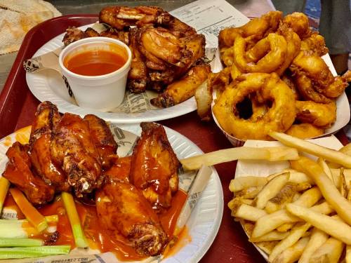 Two plates of chicken wings with sauce, onion rings, and fries on a red tray. Dipping sauce in a cup.