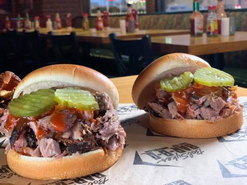 Two pulled pork sandwiches with pickles and barbecue sauce on soft buns, served on a paper-lined table in a restaurant.