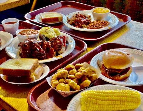 A spread of barbecue dishes on trays, including ribs, baked beans, cornbread, and a sandwich, with corn on the cob.
