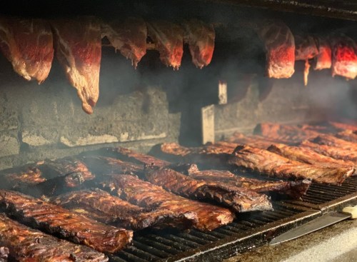 A smoky barbecue grill filled with various cuts of meat, including ribs and hanging pieces, cooking to perfection.