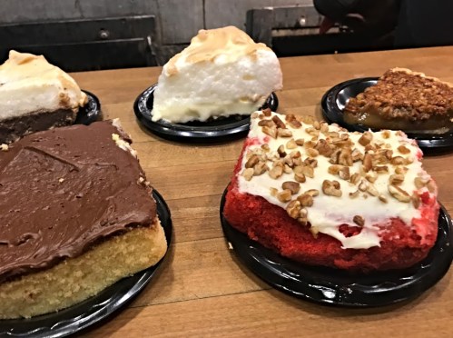 A variety of cakes on black plates, including chocolate, red velvet, and cream pie, displayed on a wooden table.