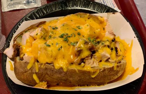 A loaded baked potato topped with shredded cheese, chopped chicken, and green onions, served in a black tray.