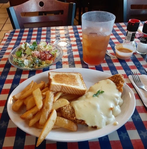 A plate of fried fish topped with cheese, served with fries, toast, a side salad, and a glass of iced tea.