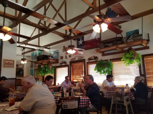 A bustling restaurant interior with wooden beams, fans, and diners enjoying meals at checkered tables.