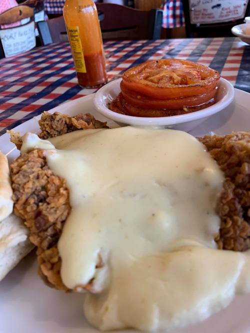 A plate of fried chicken smothered in creamy sauce, served with biscuits and a side of sliced tomatoes.