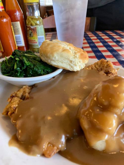 A plate of fried chicken smothered in gravy, served with collard greens, a biscuit, and hot sauce on the side.
