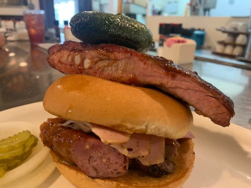 A stacked sandwich featuring a bun, smoked meat, and a pickle on top, served with pickles on the side.