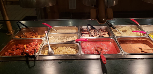 A buffet display with various dishes, including shrimp, noodles, rice, and sauces, served in metal trays with tongs.