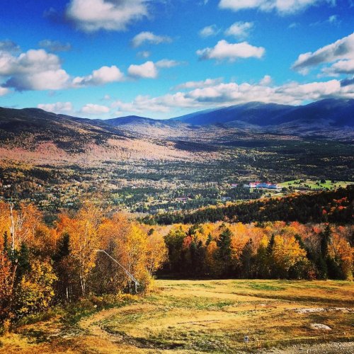 A scenic view of rolling hills and valleys, showcasing vibrant autumn foliage under a blue sky with fluffy clouds.