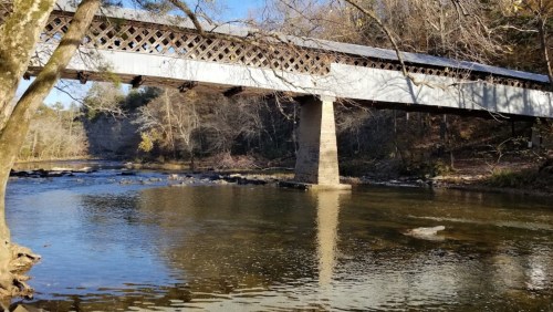 A covered bridge spans a calm river, surrounded by trees and rocky banks under a clear blue sky.