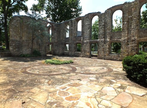 Ruins of a stone structure with arched windows, surrounded by greenery and a stone pathway in a sunny setting.