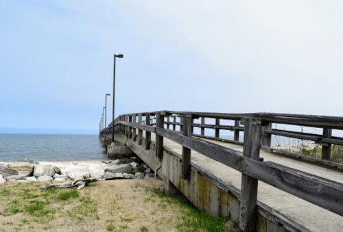 A wooden pier extends over calm water, with grassy areas and rocks along the shore under a cloudy sky.