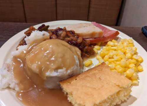 A plate of food featuring mashed potatoes with gravy, baked beans, ham, corn, and a slice of cornbread.