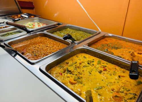 A variety of colorful curries and dishes in metal trays on a buffet table, with vegetables and rice options visible.