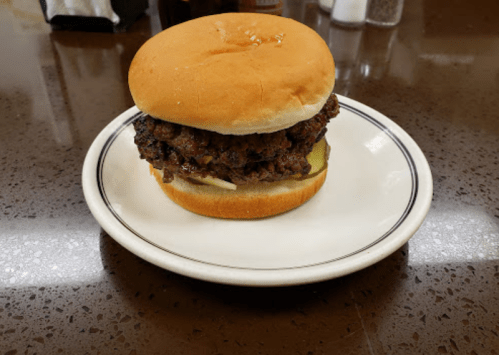 A juicy hamburger with pickles on a bun, served on a white plate on a dark countertop.