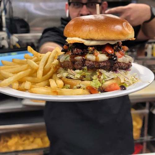 A person holds a plate with a large burger topped with vegetables and sauce, accompanied by a side of French fries.