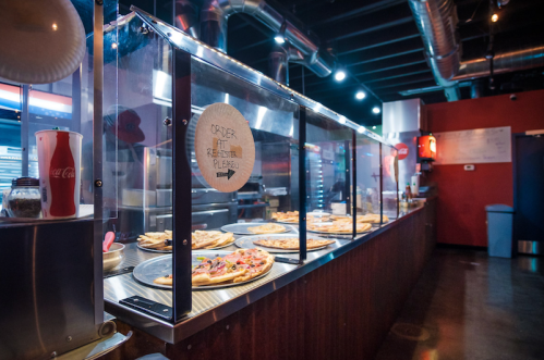 A pizza counter with various pizzas displayed under glass, featuring a sign that says "Order at Register Please."
