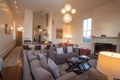 Cozy living room with modern decor, large windows, and a kitchen area, featuring comfortable sofas and stylish lighting.
