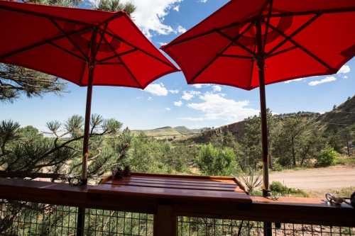 A scenic view from a deck featuring two red umbrellas, overlooking green hills and a blue sky with clouds.